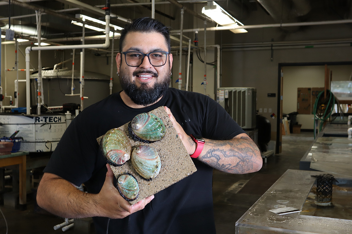 A student with a beard, glasses and tattoos holds up a block of cement with four living abalone shells on it